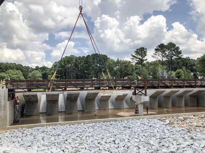 The last bridge span was positioned on the spillway for Lake Peachtree last week. The span still has to be filled with concrete, along with other assorted work. For perspective on the scale of the structure, yes, those tiny figures at the left on a lift and at center right beneath a piling are indeed grown men. Photo/Vanessa Fleisch.