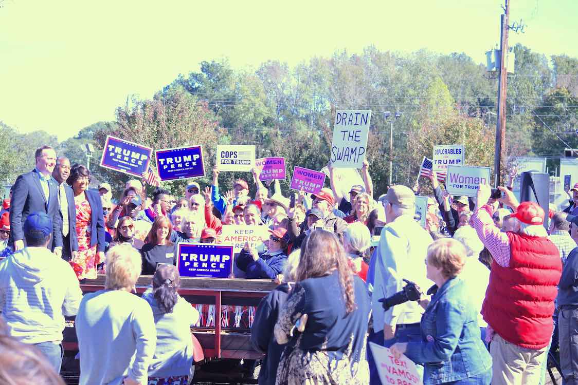 <b>Fayette native Brian Jack (L, on platform), the White House Political Director, stands with HUD Secretary Dr. Ben Carson and Carson's wife waving at the Fayetteville Trump rally supporters. Photo/Cal Beverly.</b>